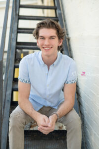 A senior boy is wearing a light blue shirt with khakis pants is sitting on a set of stairs with his arms leaning over his legs and smiling at Khim Higgins Photography.