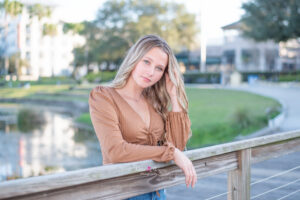 A senior blond girl is wearing a tan long sleeve shirt is leaning over a rail by the lake.