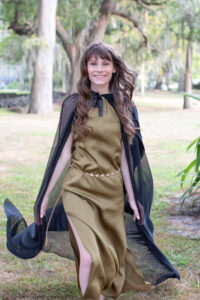 senior girl in a long green dress is wearing a black cape walking towards Orlando Senior Photographer smiling.