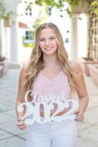 A senior girl in a pink top and white pants is holding a sign "class of 2022" as Khim Higgins Photography takes her picture.