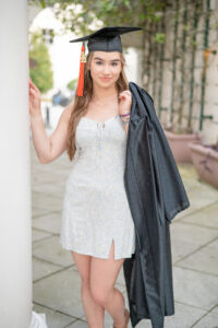 Mary, a senior at Oviedo High School is wearing her cap and holding her gown over her shoulder. Mary is wearing a short white dress and smiling at Khim Higgins Photography.