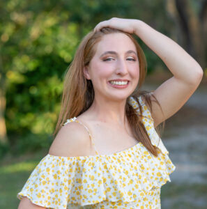 A senior girl from Oviedo High School is wearing a short yellow dress, with one hand one brushing her hair back and the other hand on her hips is smiling beautifully at Khim Higgins Photography.