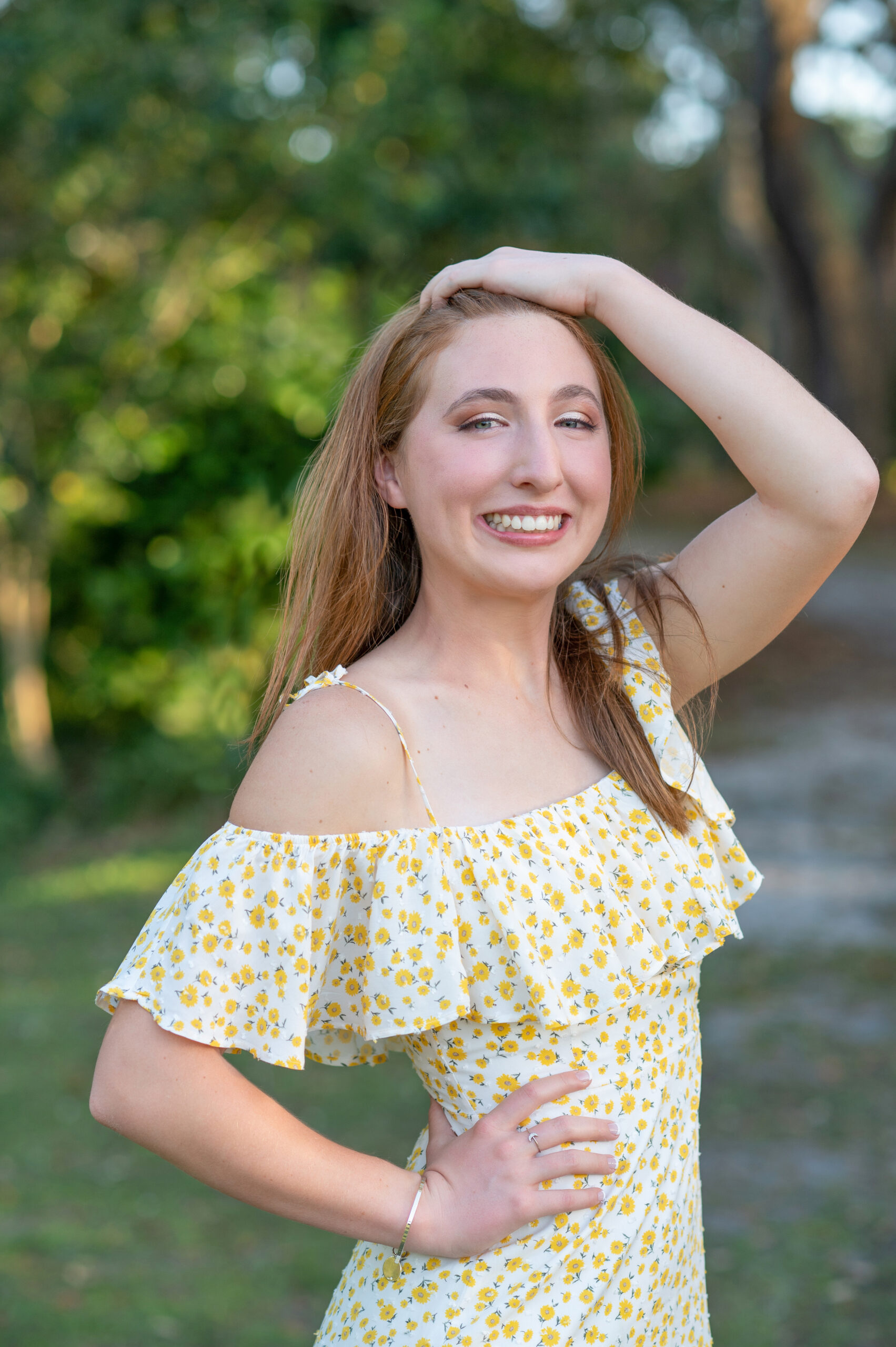 A senior girl from Oviedo High School is wearing a short yellow dress, with one hand one brushing her hair back and the other hand on her hips is smiling beautifully at Khim Higgins Photography.