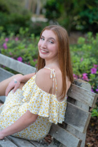 An Oviedo High School senior girl in a yellow dress is sitting on a bench and looking behind her over her left shoulder smiling at Orlando senior photographer, Khim Higgins.