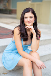 Winter Park High School senior girl in a blue dress is sitting on some stairs with one hand on her chin looking at Khim Higgins Photographer.