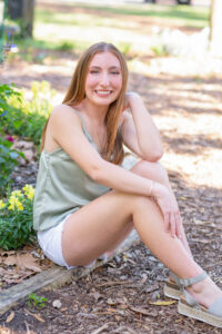 Sara is a senior girl from Oviedo high school and she is wearing a sage top and white shorts. Sara is sitting on a piece of wood and smiling at Orlando Senior photographer, Khim Higgins.