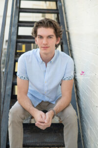 senior boy aiden is wearing a light blue shirt and khaki pants and he is sitting on a set of black stairs leaning over his knees looking at the camera with a serious face.