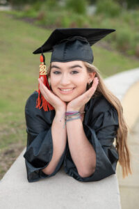 OHS senior graduation photo shoot with Khim Higgins Photography. Mary is wearing her black cap and gown and laying on her belly on the sidewalk with her hands on her face smiling at Orlando Senior PHotographer.