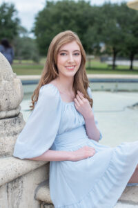 A lake mary senior girl is wearing a blue dress and sitting by a fountain.