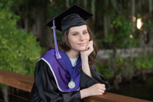 Timber Creek senior girl is wearing her black and purple cap and gown is leaning on a dock by the lake as Orlando senior photographer takes her picture.