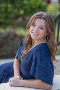 senior girl in a blue dress is sitting on stairs leaning back on one arm and looking over her left shoulder