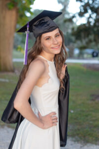 Timber Creek senior girl is wearing a white dress, with her graduation cap on her head and holding her graduation gown looking at Orlando senior photographer.