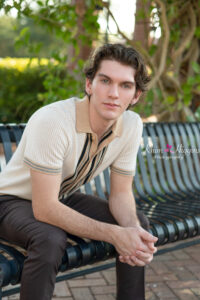 Orlando senior boy photo shoot with Aiden and he is wearing a cream color short sleeve sweater with dark brown slacks and sitting on a bench looking sideway at the the camera.