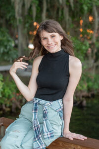 Timber Creek senior girl is wearing a black sleeveless top and jeans sitting at the dock playing with her hair smiling at Orlando senior photographer, Khim Higgins.