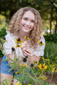 A senior girl wearing a white top and jean shorts is holding sunflowers in her hands while smiling at Khim Higgins Photography.