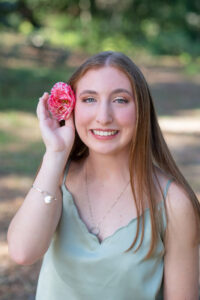 Sara has a pink rose on the side of her right ear as she cups it with her right now. She is smiling and sitting on the wood ledge as Khim Higgins Photography takes her photo. 