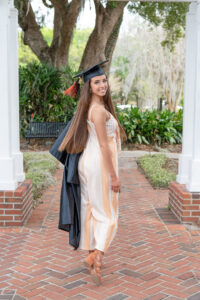 Senior girl wearing her graduation cap and gown is looking over her shoulder at the camera as Orlando Senior Photographer takes her shot.