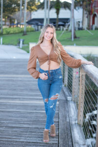 A Hagerty High School Senior girl is wearing a long sleeve shirt that ties in the front with jeans is standing on a pier by the lake.