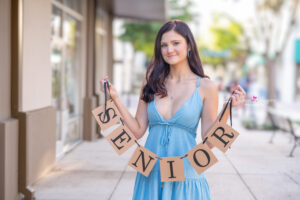 Winter Park High School senior is wearing a blue dress holding a 'senior' banner smiling at Orlando Senior Photographer.