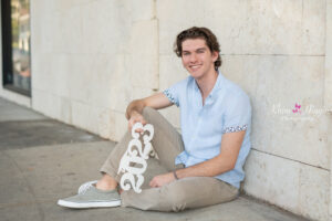 A senior boy from Winter Park High school is wearing a light blue shirt and khaki pants is sitting on a sidewalk leaning on a wall and he is holding a sign that says 2023.