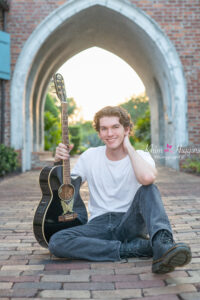 A Winter Park High School senior boy is wearing a white t-shirt and blue jeans is sitting on a walkway and holding his guitar.