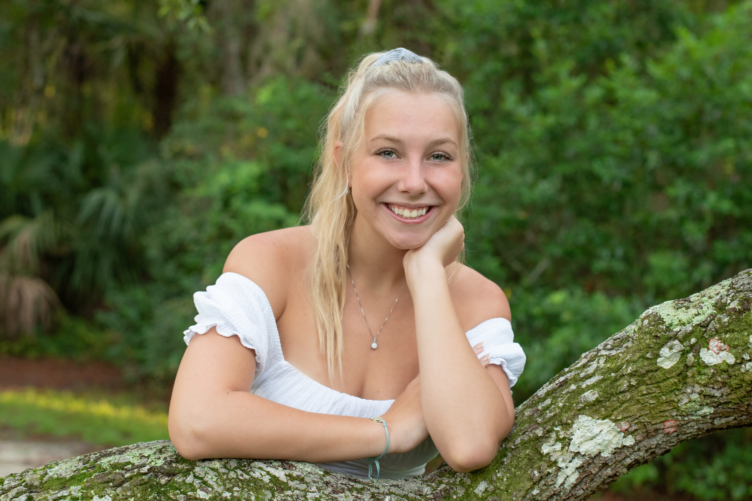 A Lake Mary High School senior girl photo shoot with Khim Higgins Photography. The senior girl is wearing a white top with blue jeans and she is leaning on a tree branch.