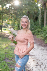 Senior girl photo shoot in Lake Mary, Florida of a blond girl wearing a pink top and blue jeans standing and holding a vine in one hand smiling at Khim Higgins Photography.