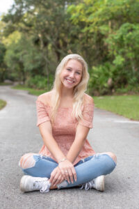 Lake Mary senior photo shoot with a blond girl wearing a pink top and blue jeans. Senior girl is sitting on the road smiling at Khim Higgins photography.