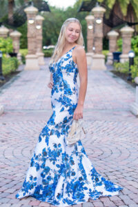 Senior girl in her blue and white long prom dress is holding her shoes in one hand and she is looking over her left shoulder as Khim Higgins Photography takes her photo.