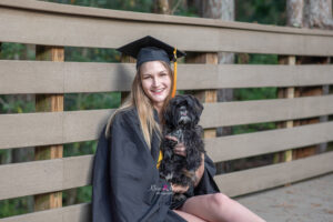 a ucf grad wearing her cap and gown is holding her little black dog and sitting on a boardwalk.