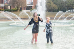 ucf graduate is standing in the reflection pond with her grandmother and they are both drinking champagne from the bottle.