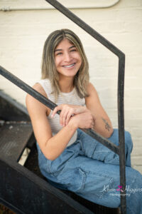 senior girl in a cream color top and blue jeans is sitting on a set of black iron staircase.