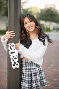 orlando senior girl in a white sweater and checkered skirt is holding a 2023 sign leaning on a pole.