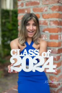 senior girl in a blue prom dress is holding a 'class of 2024 sign.'