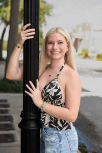 A blond Hagerty High School senior girl wearing jeans and a black and white top is holding onto a light pole on a sidewalk.