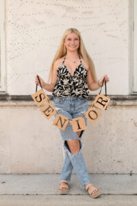 A blond Hagerty High School senior girl wearing jeans and a black and white top is holding a 'senior' sign.