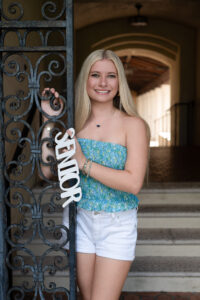 A blond Hagerty High School senior girl wearing white shorts and a blue top is standing in front of an archway at Rollins College holding a 'senior' sign.