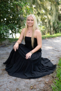 A blond Hagerty High School senior girl in a black sundress is sitting by a lake.