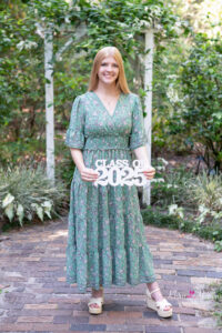 Oviedo high school senior girl is wearing a long green dress standing in a garden holding a 'class of 2025' sign.