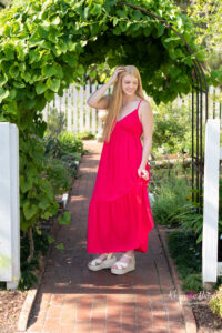 Oviedo high school senior in a long, red sun dress is standing in a garden.