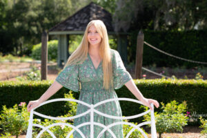 Oviedo high school senior girl is wearing a long green dress standing behind a white metal bench at Leu Gardens.