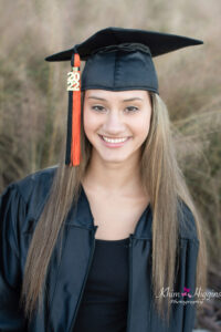 A senior girl wearing her black cap and gown is smiling at the camera in a close headshot.