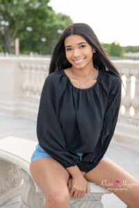 A senior girl wearing a black long sleeve blouse and wearing jean shorts is sitting on a bench.