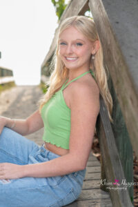 A blond senior girl wearing a green tank top and jeans is sitting on the boardwalk at the beach.