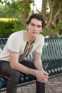 A senior boy wearing a cream color knit sweater and brown dress pants is sitting on a bench and leaning over with his elbows on his knees.