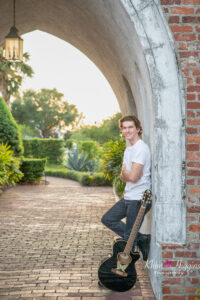 A senior boy wearing a white t-shirt and black jeans is leaning against a brick wall with his guitar next to his leg.