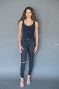 a beautiful brunette girl is wearing a black tank top and black jeans is standing in front of a gray backdrop for her modeling photo with khim higgins photography.