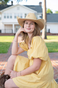 a senior girl in a yellow dress and cowboy hat is sitting on a step happily looking at a lake mary photographer.