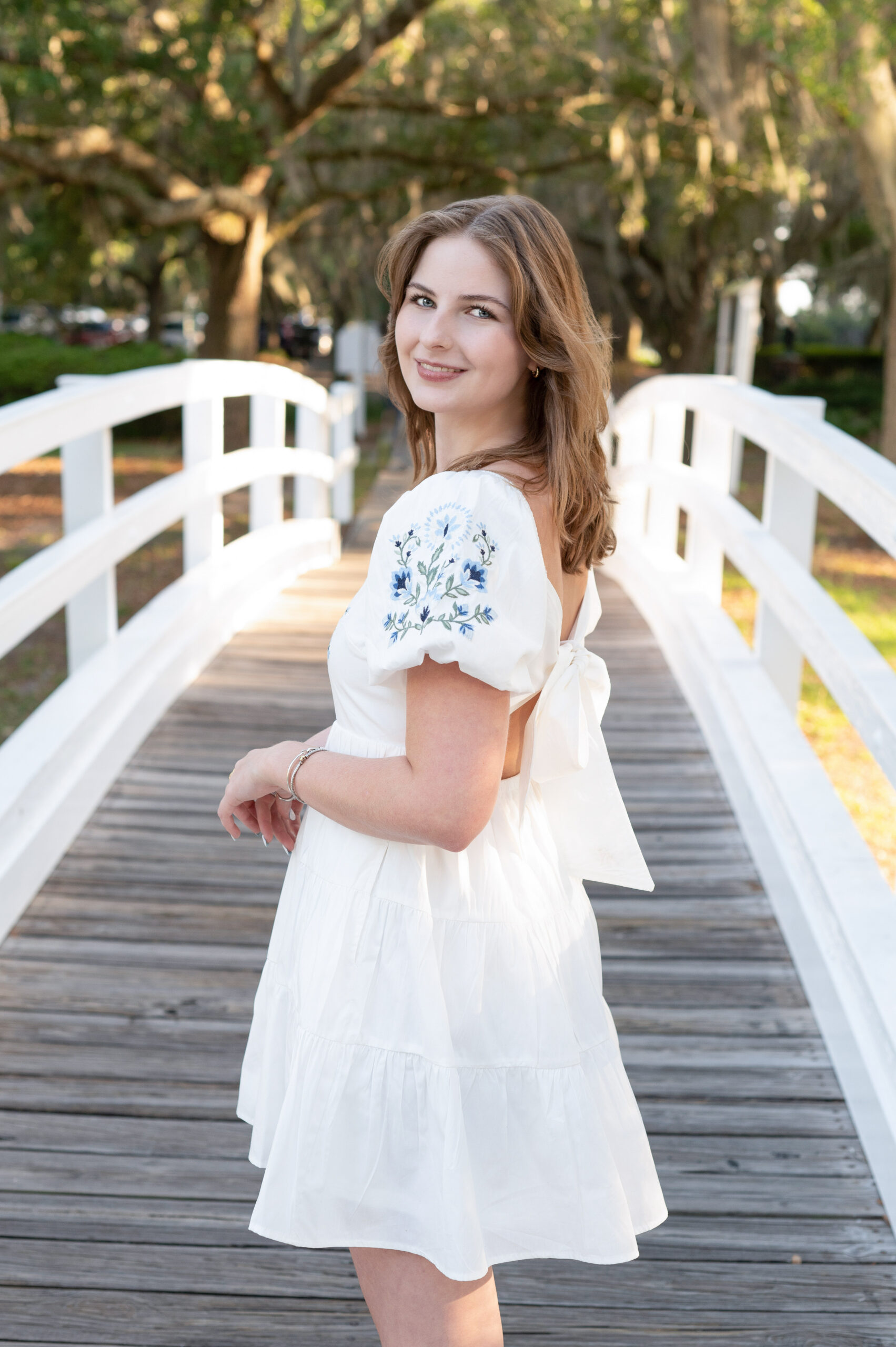 Lake Mary senior photography session featuring a high school girl in a white floral dress, smiling while standing on a wooden bridge surrounded by greenery.
