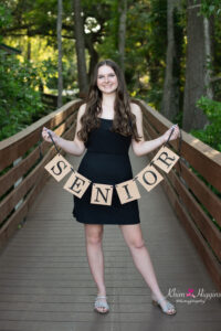 oviedo senior girl in a black dress is holding a 'senior' banner standing on a dock at lake mills with Khim higgins photography.
